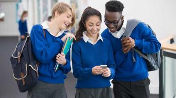 Three smiling students wearing blue school uniforms with mobile phone in school corridor